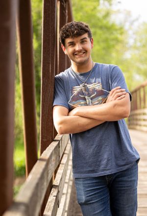 Person wearing a graphic t-shirt stands with arms crossed on a wooden bridge in a natural setting on a sunny day.
