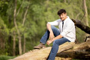 A person in jeans and white button-up shirt relaxes casually on a fallen tree trunk in a wooded setting.