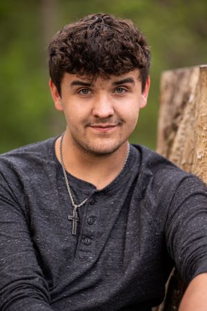 A person in a dark gray henley shirt sits outdoors against a tree trunk with a soft natural background.