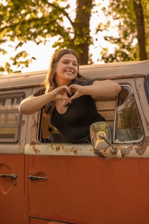 A vintage orange Volkswagen van with someone smiling and making a heart shape through the window in golden sunlight.