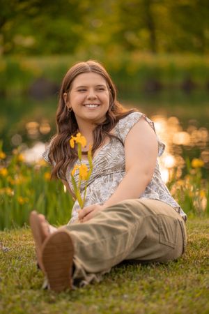 A person in casual attire sits on grass holding a yellow flower during golden hour with bokeh lighting in background.