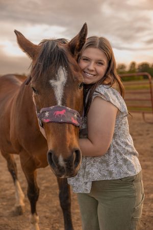 A brown horse with a colorful halter being embraced warmly in an outdoor paddock at sunset.