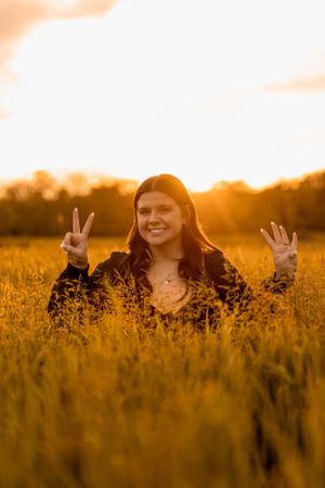Person making peace signs while standing in golden field at sunset with warm glowing backlight.