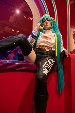 Cosplay outfit featuring long turquoise hair, retro-style t-shirt, and black shorts against a red backdrop with dramatic lighting.
