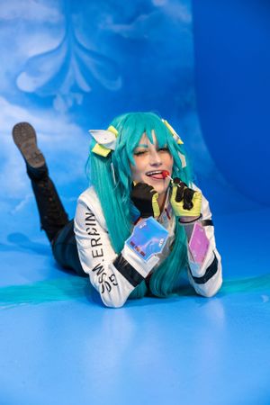 Cosplayer wearing turquoise wig and cyberpunk-inspired costume lying on blue background holding a microphone.
