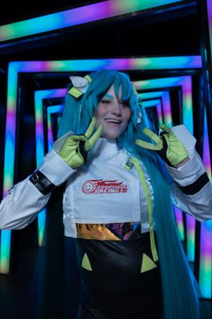 Cosplay photo showing a figure in turquoise wig and racing-style outfit posing against a colorful illuminated geometric background.