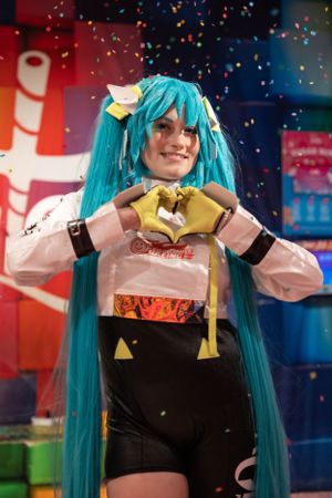 Cosplayer wearing a turquoise wig poses in costume with confetti falling at a convention event.