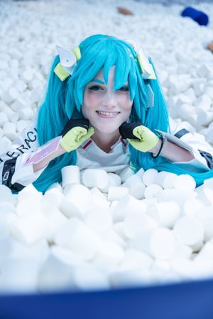 A cosplayer with bright turquoise twin tails and yellow gloves poses playfully amidst white plush objects.