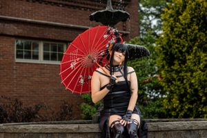 Cosplay fashion model in gothic style dress poses with traditional red Japanese parasol against brick building exterior.