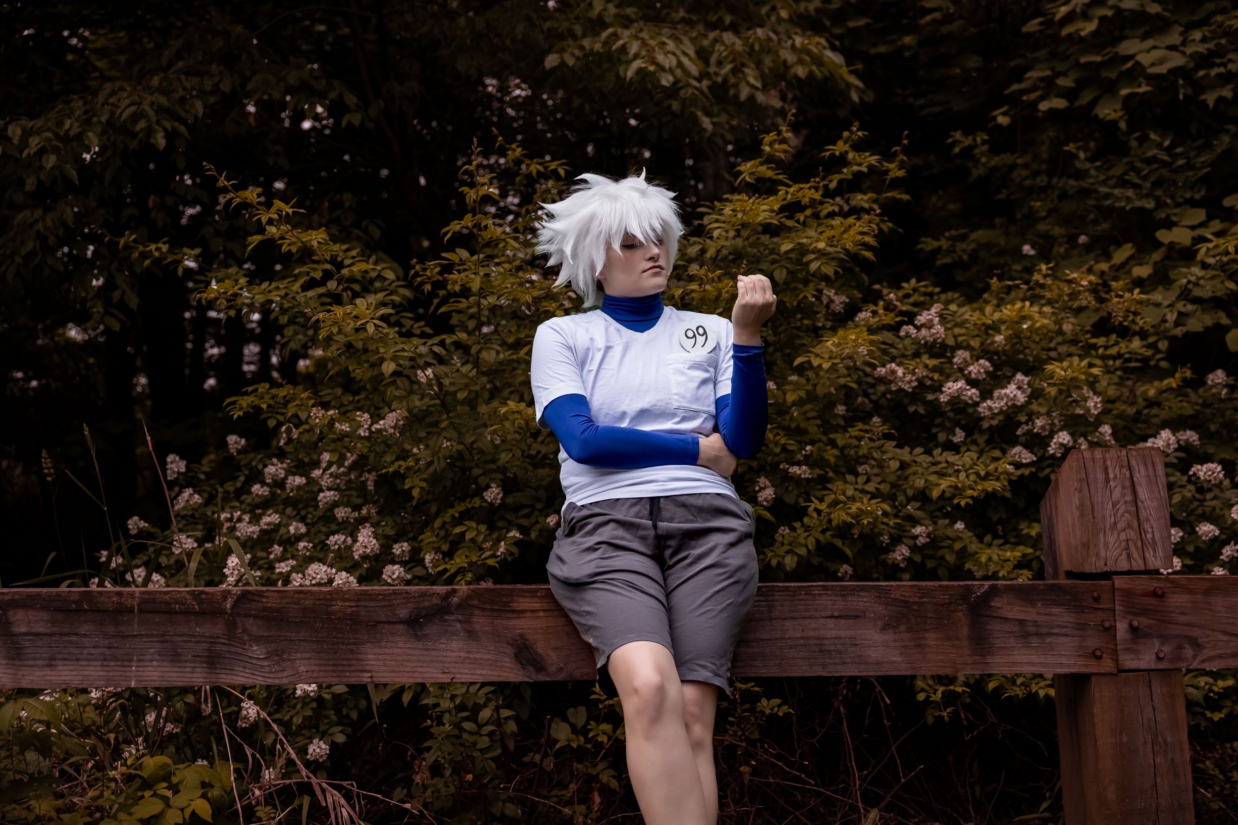 An anime cosplayer in a blue and white top with grey shorts poses on a wooden railing against autumn foliage.