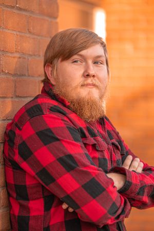 Person in red and black buffalo plaid flannel shirt poses with arms crossed against brick wall in warm lighting.