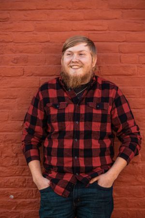 Smiling person wearing a red and black buffalo plaid flannel shirt stands against a brick wall.