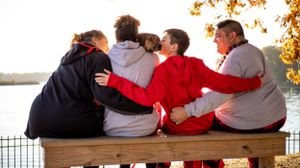 A group sits together on a wooden bench overlooking a lake during autumn with their arms around each other.