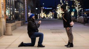 A romantic nighttime proposal scene on an illuminated city street with decorative lights and trees.