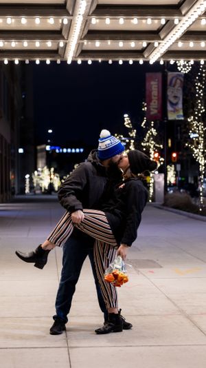 A couple shares a romantic dip under festive holiday lights on a city sidewalk while holding a flower bouquet.