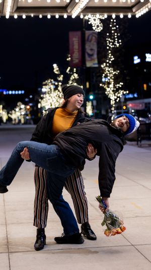 Two people perform a playful dip pose on an illuminated city street at night with decorative lights in background.