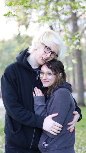 A couple in dark hoodies sharing an affectionate embrace outdoors on a sunny spring day among blossoming trees.