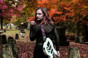Gothic style person poses in a cemetery with a guitar during autumn surrounded by vibrant orange and red foliage.