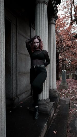 Moody gothic fashion photo with dark clothing against stone columns in autumn cemetery setting with pink leaves.