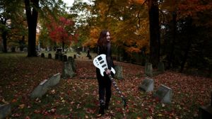 A spooky Halloween skeleton figure stands among fallen autumn leaves and gravestones in a cemetery.
