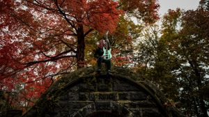 A person stands atop a stone bridge arch surrounded by vibrant red autumn foliage in a forest.
