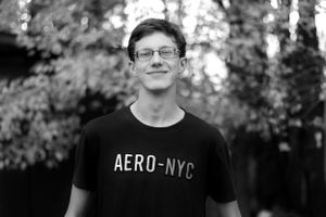 Black and white portrait of someone wearing an Aero-NYC t-shirt smiling outdoors with trees in the background.