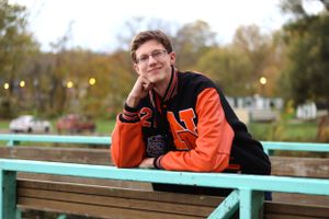 College student in orange varsity jacket leans on turquoise railing outdoors during autumn.