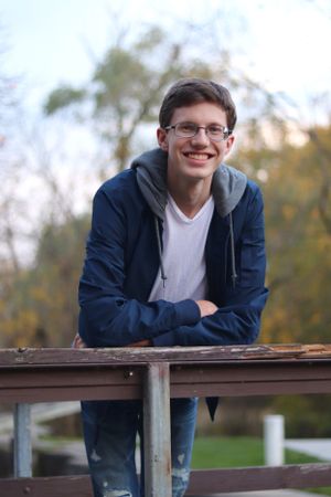Casual portrait showing someone in glasses and a navy jacket leaning on a wooden railing outdoors during autumn.