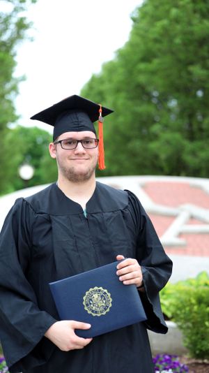 A graduating student in cap and gown holds a diploma while posing outside on a sunny day with greenery in the background.