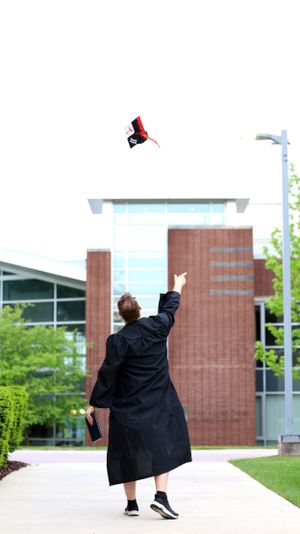 Graduate in black cap and gown throws their graduation cap in the air on campus walkway during celebration.