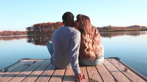 Two people sitting together on wooden dock by tranquil lake during sunset with trees along shoreline.