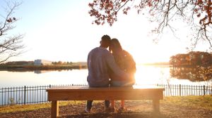 A couple sits on a wooden bench overlooking a serene lake at sunset with autumn trees framing the romantic scene.