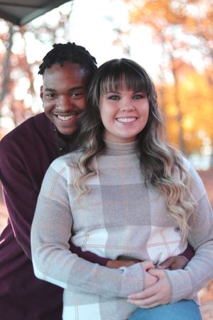 A couple embraces and smiles at the camera while wearing cozy sweaters on an autumn day with warm bokeh lighting in background.