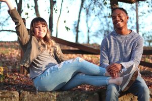 Two people sitting together on fallen logs in a forest on an autumn day with scattered leaves on the ground.