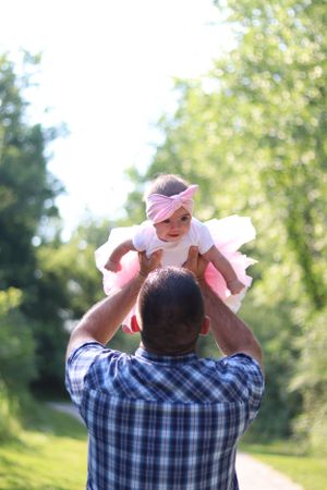 A person in a plaid shirt lifts a baby wearing white and pink outdoors in a sunny park setting.