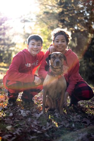 Two people in red hoodies pose outdoors with their brown dog among autumn leaves during golden hour sunset.