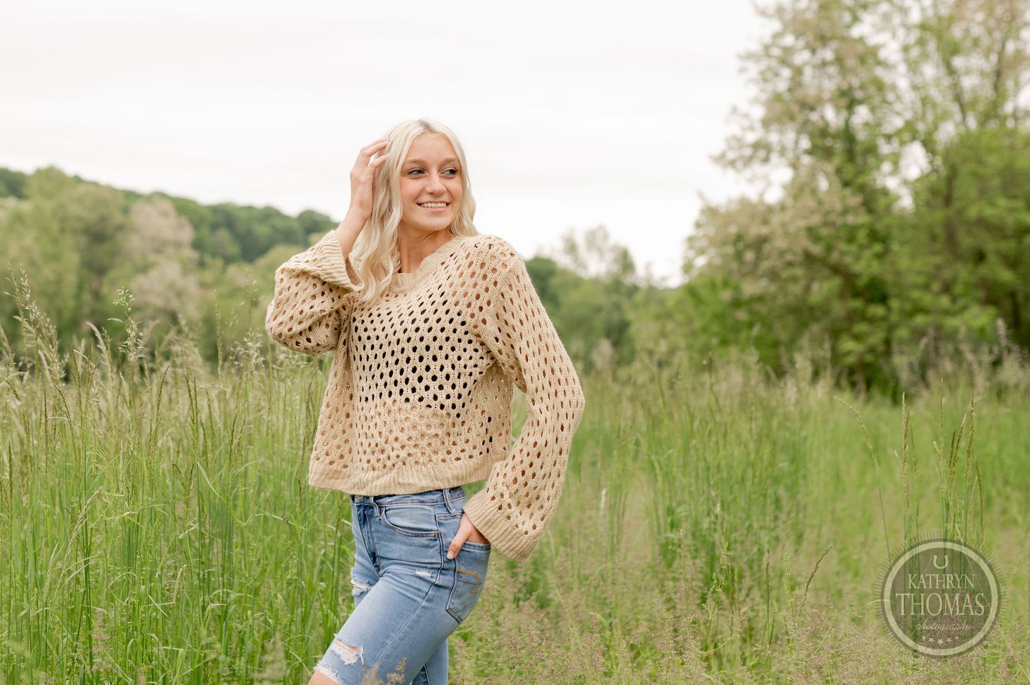 Trendy boho style featuring a cream openwork sweater and ripped denim in a grassy field setting.