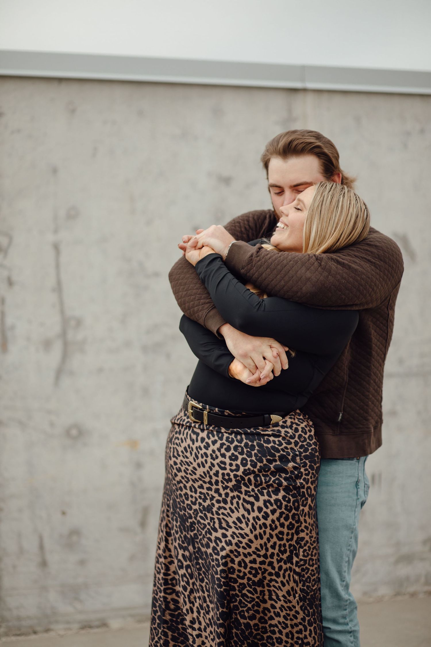 Couple embraces tenderly against concrete wall backdrop, person wearing brown sweater and jeans hugging person in leopard print skirt.