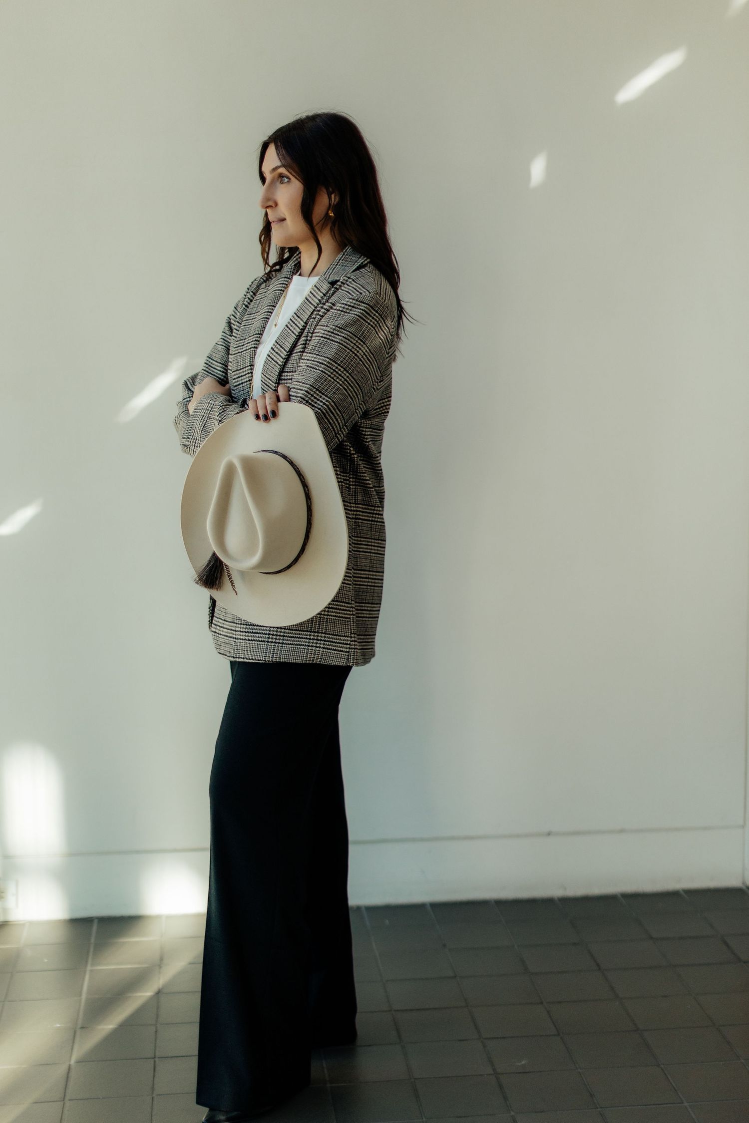 Woman in cozy gray cardigan and maxi skirt holds cream fedora hat while standing against white wall in natural light.