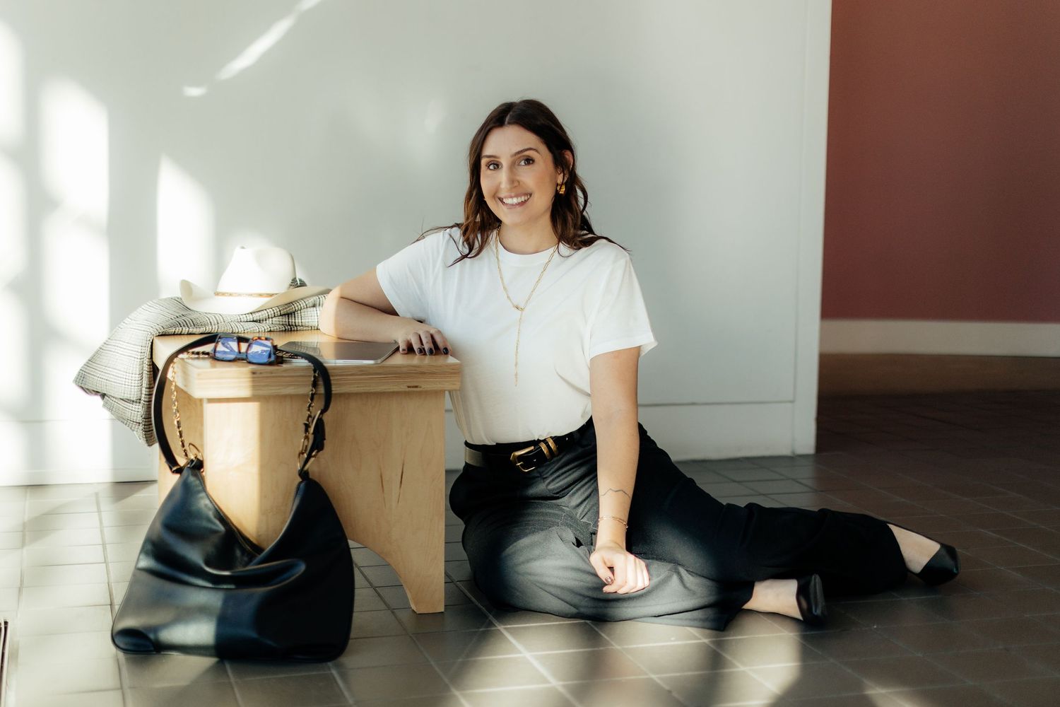 Someone in a white t-shirt and black pants sits casually on a wooden floor next to a modern drum set.