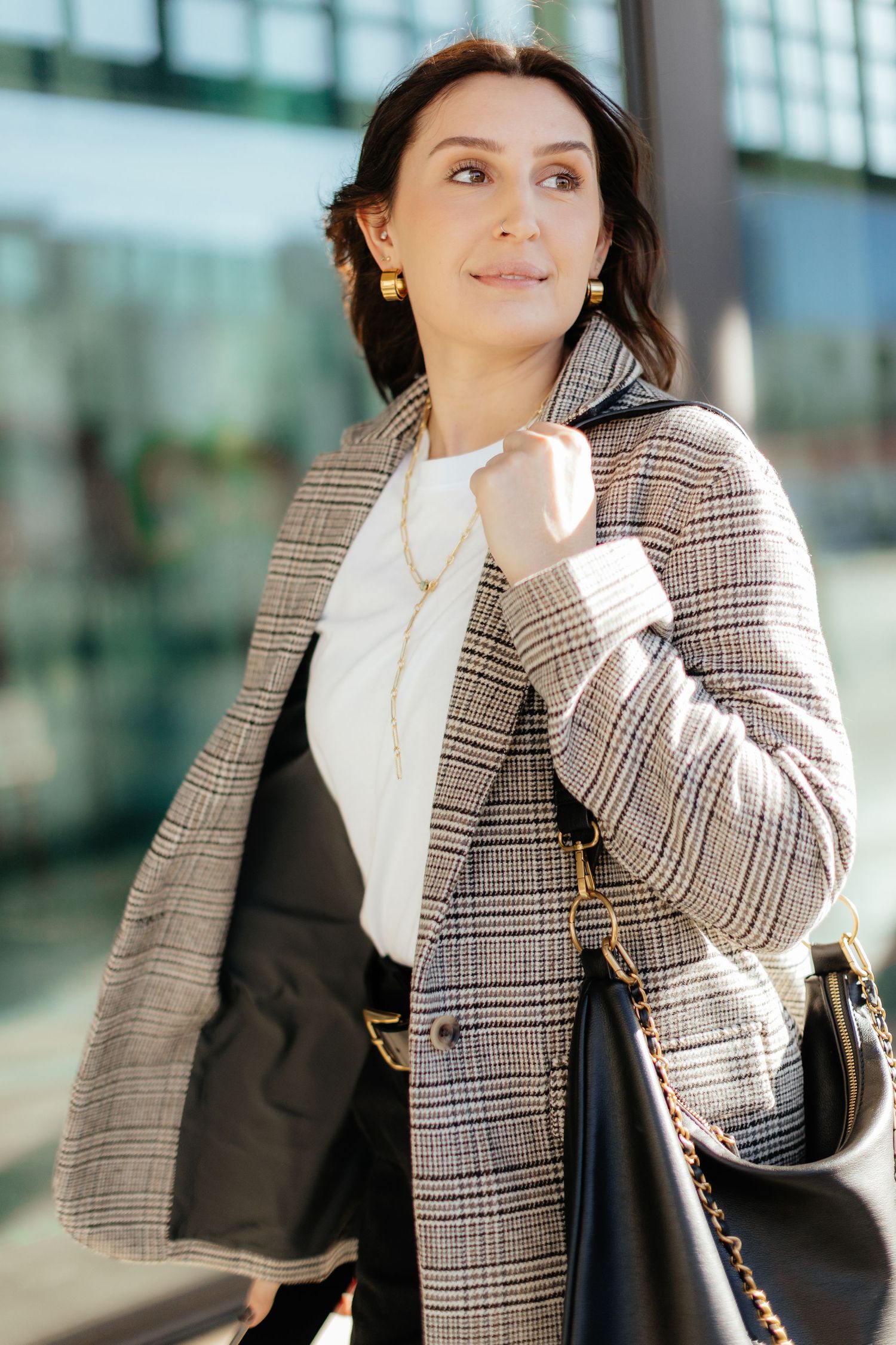 Professional woman in plaid blazer and white blouse carrying black handbag against modern glass building exterior.