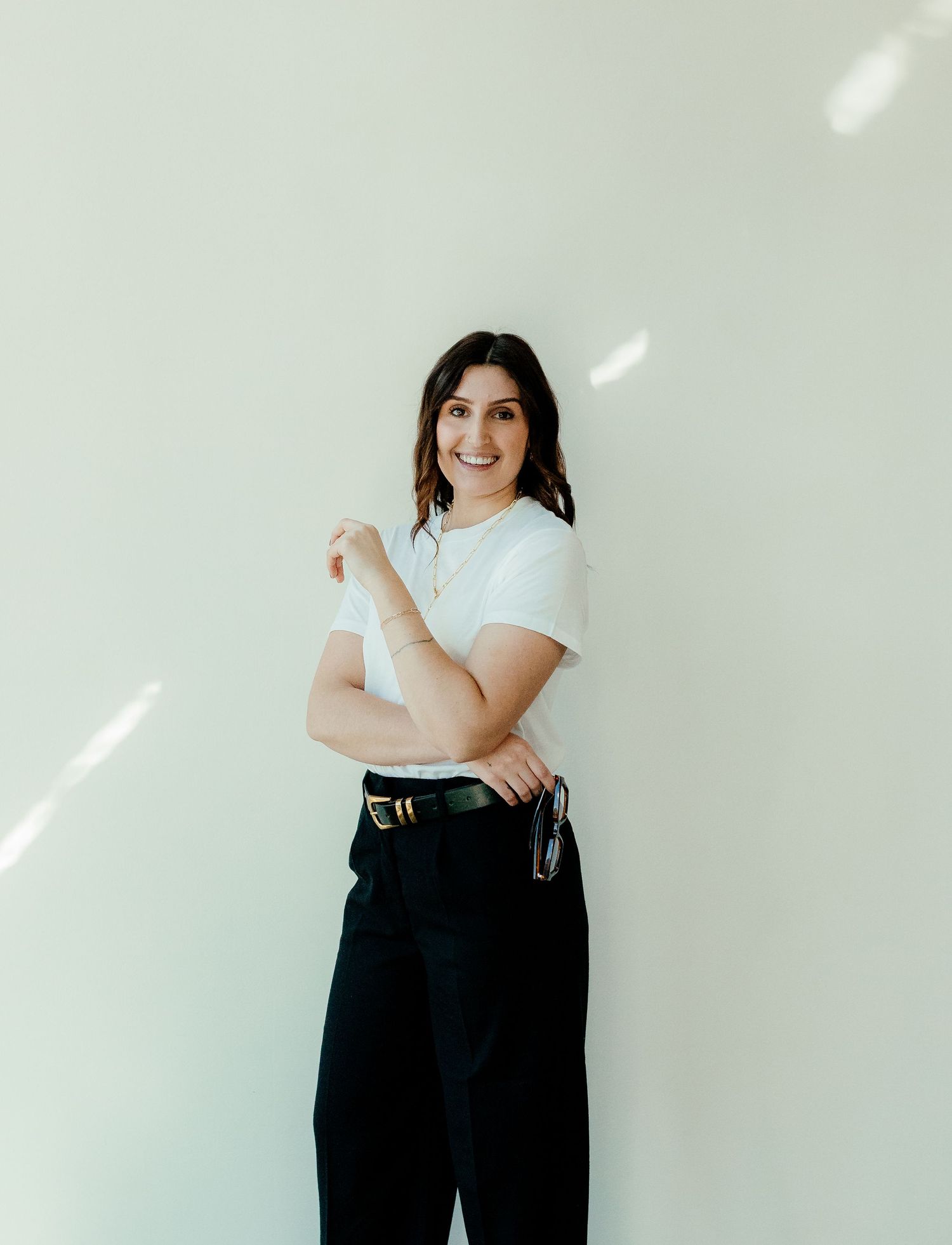 A person in a white t-shirt and black trousers stands against a light wall with sun rays casting diagonal shadows.