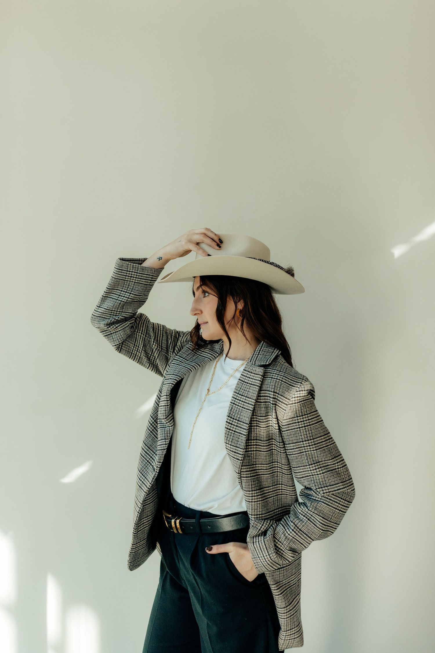 Fashion portrait of someone wearing a plaid blazer, white hat, and dark pants against a light wall.