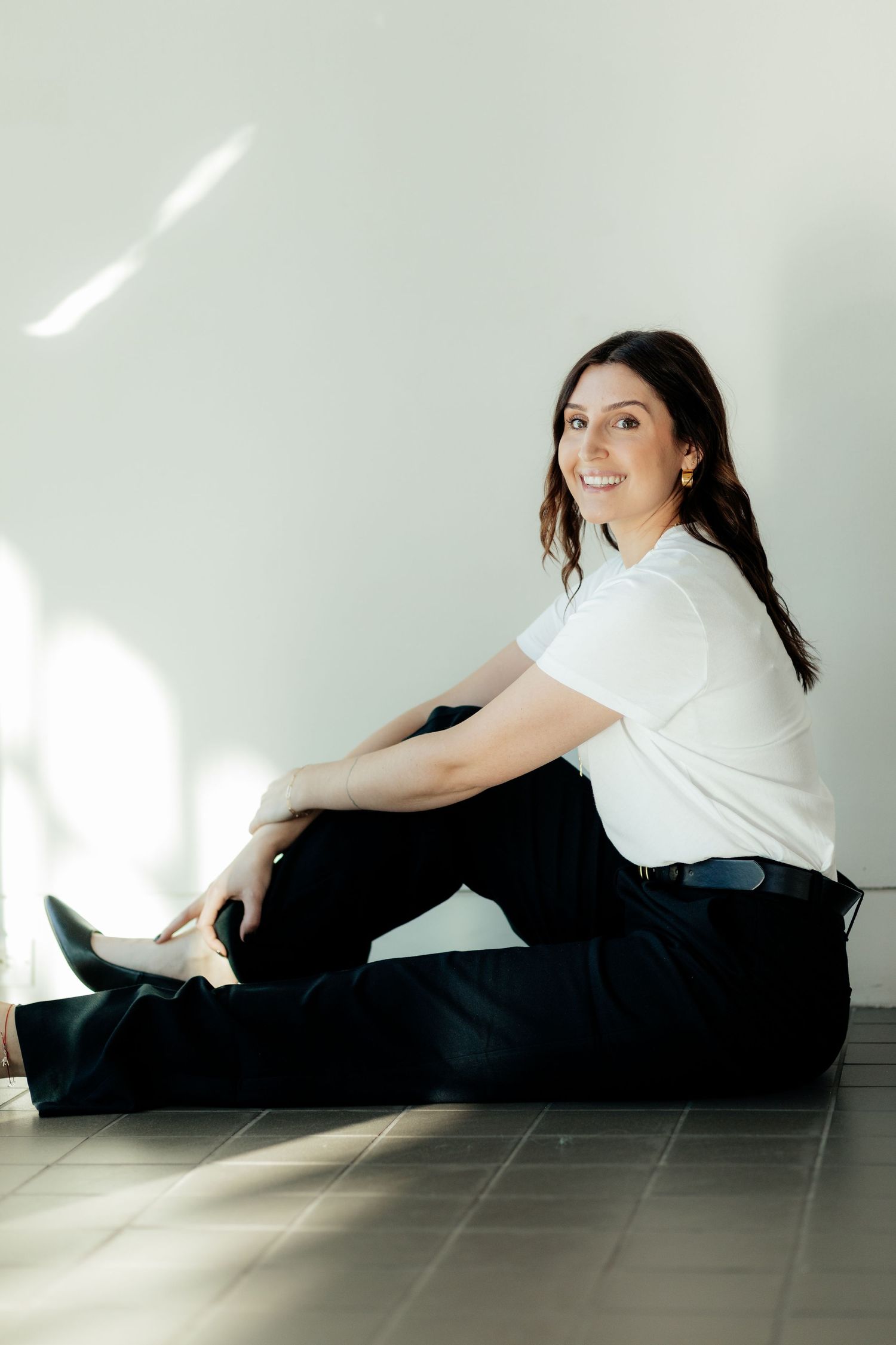 Woman in casual white top and dark pants sits on floor near window with soft natural lighting creating shadow patterns.