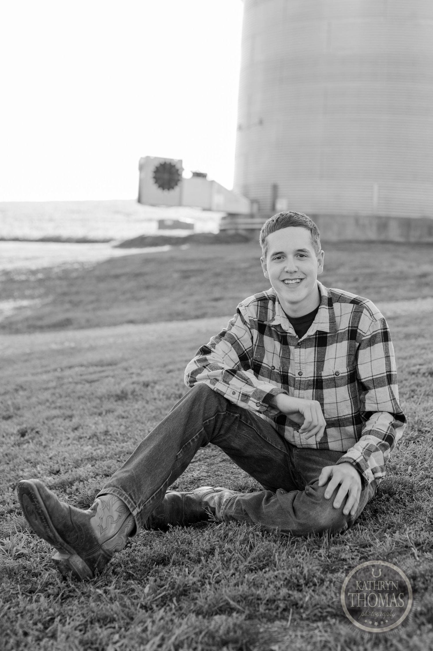A casual outdoor portrait by a grain silo with someone wearing a plaid flannel shirt sitting on the grass.