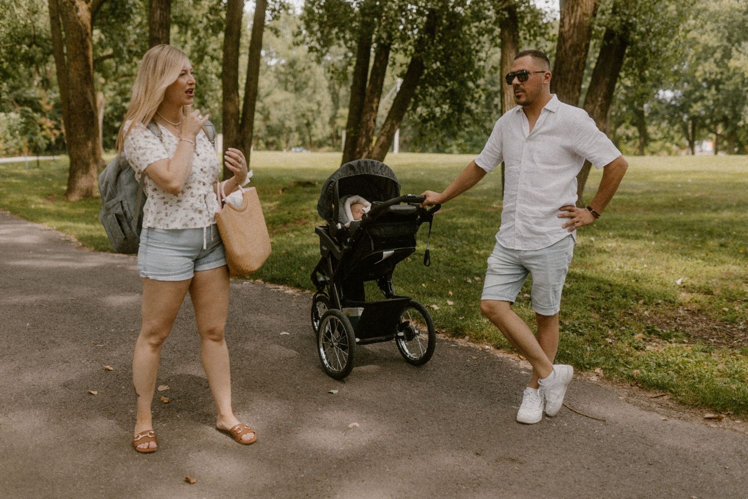 A couple walks together with their stroller on a tree-lined path in a park.