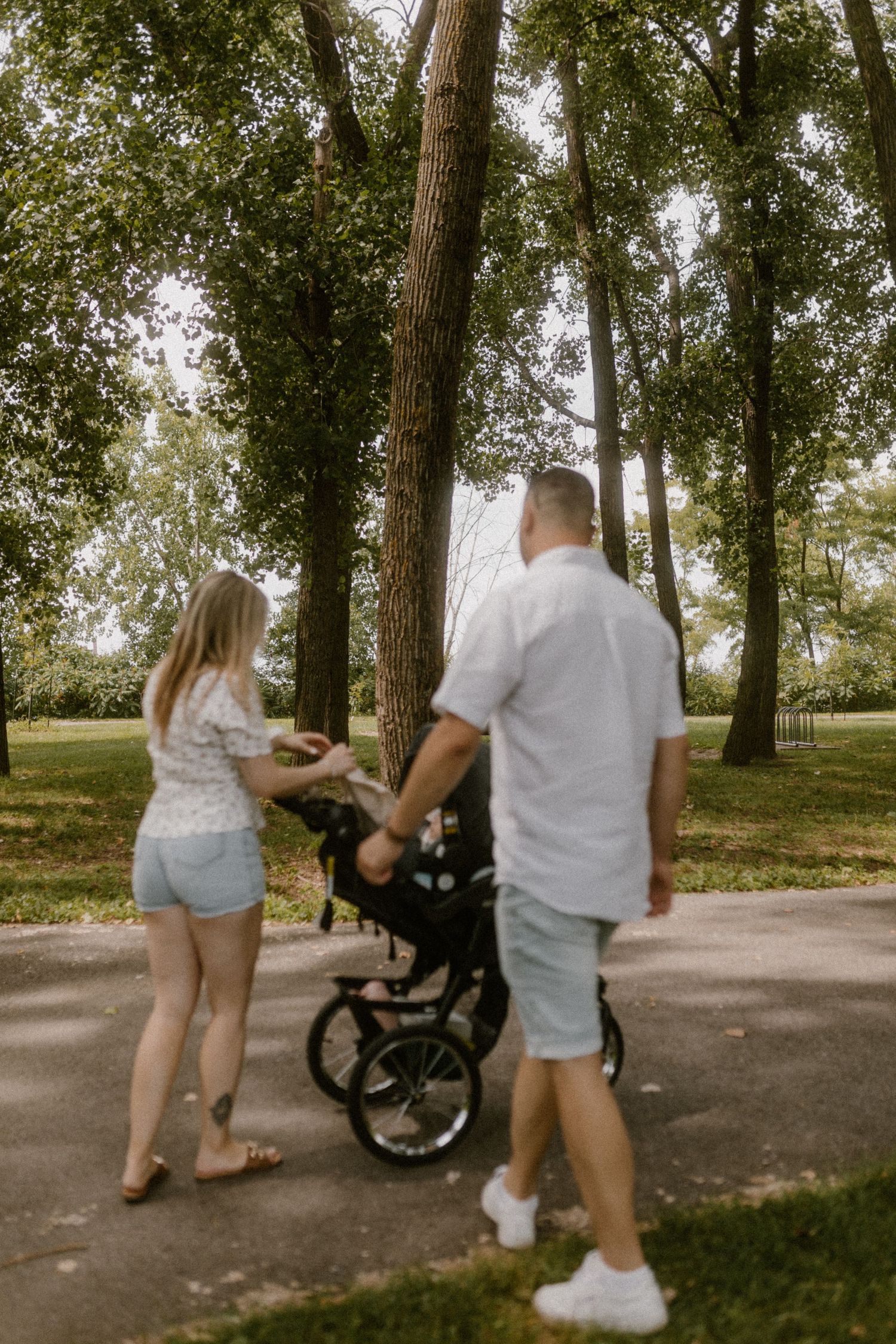 A couple walks with their stroller through a wooded park path on a sunny afternoon.