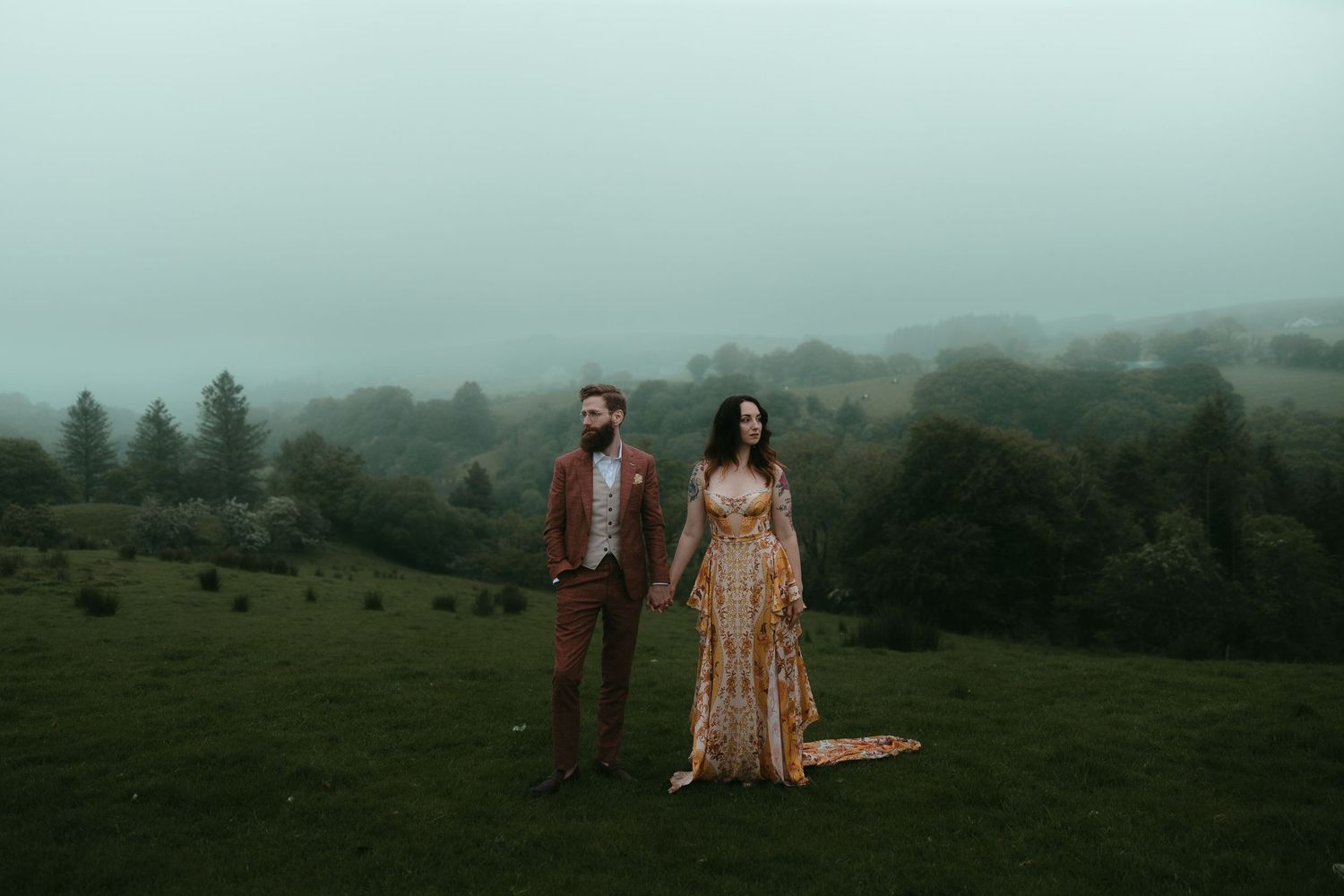A couple stands in a misty green field wearing wedding attire for an atmospheric outdoor portrait.