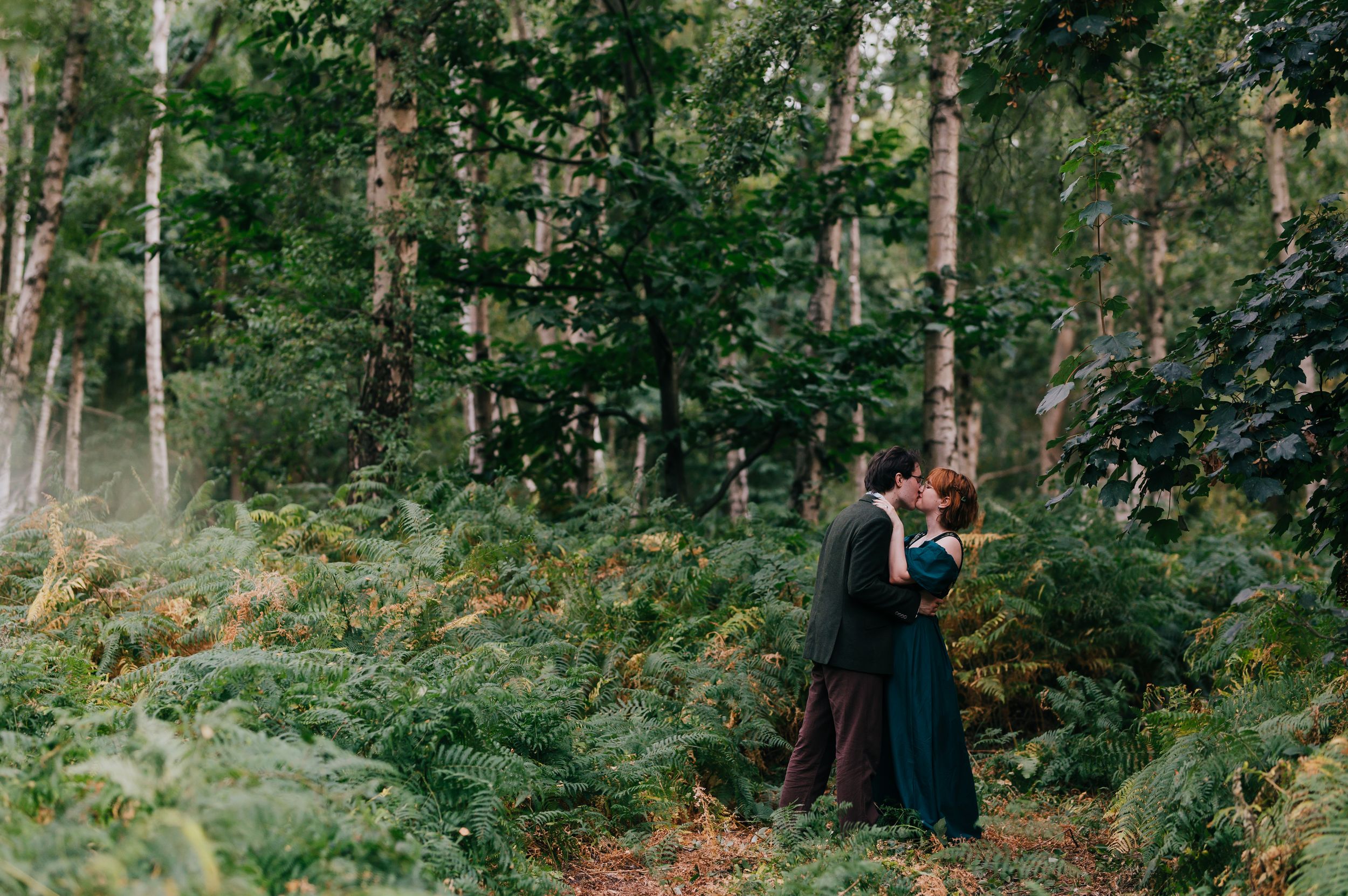 Couple sharing a romantic kiss surrounded by lush green ferns and tall trees in a misty forest setting.