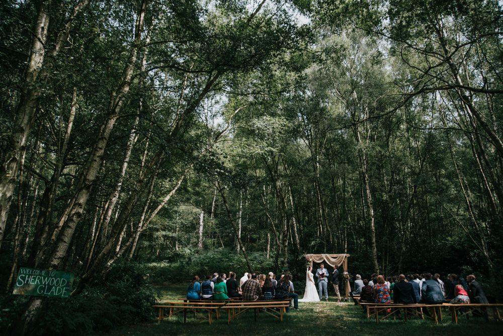Outdoor wedding ceremony in a lush forest setting with wooden benches and tall trees creating a natural cathedral effect.